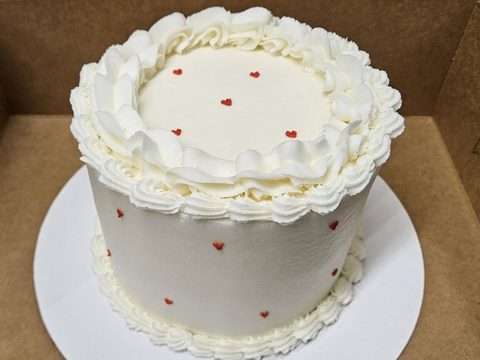 Small white frosted cake with ruffled piping and tiny red hearts, in an open bakery box