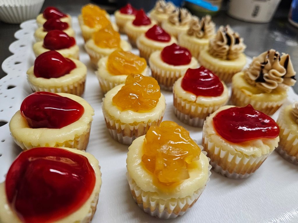 Rows of mini cheesecakes topped with cherry, orange fruit and swirled frosting